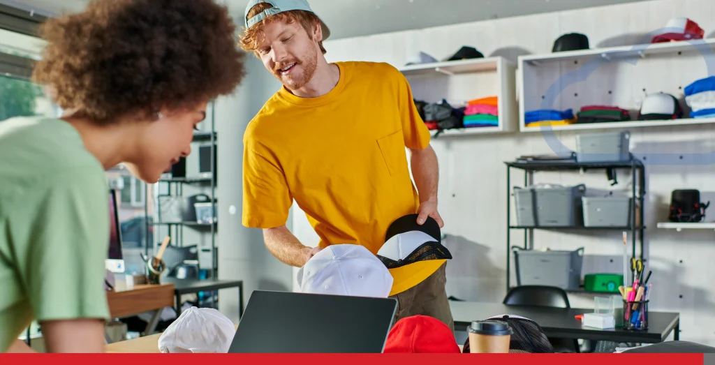 Smiling young designer holding snapbacks while talking to african american colleague using laptop Stock Photo, ft. african american & artisans - Envato